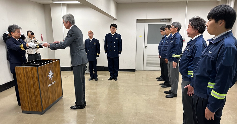 警備所での正式な儀式で、スーツ姿の男性が見物人に囲まれて海上保安庁から賞状を受け取る授賞式の様子