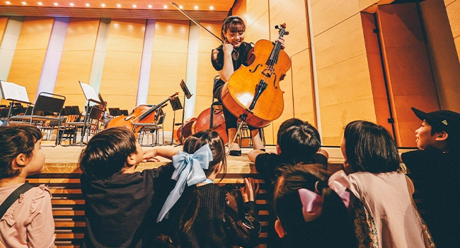 音楽家がステージ上でチェロを演奏し、子どもたちは楽器に囲まれて最前列から熱心に見入っています。